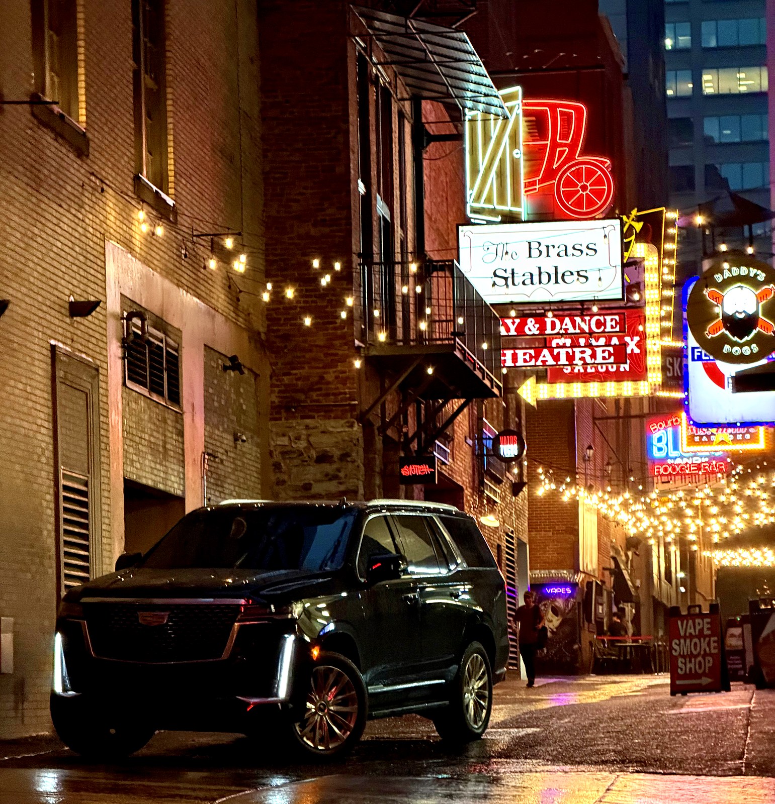 A black Cadillac Escalade parked in Printers Alley at night beneath the neon signs in downtown Nashville